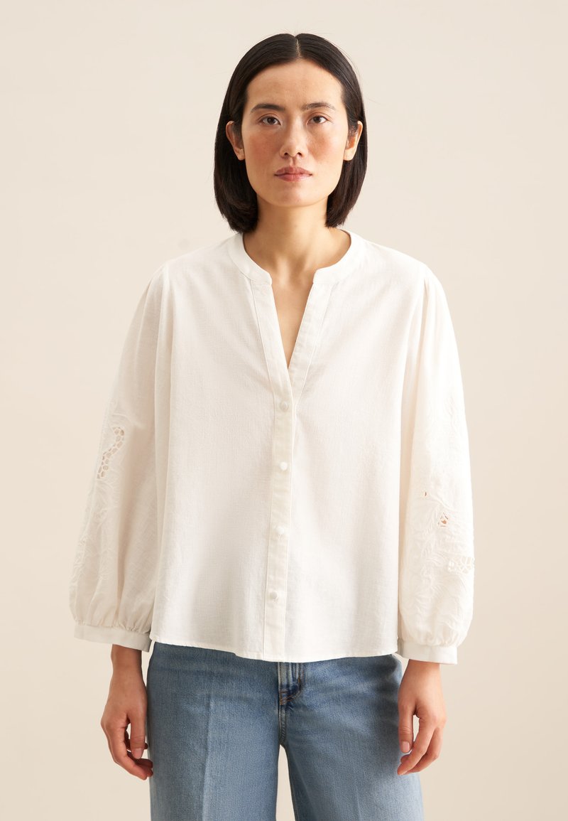 Weiße Bluse mit Knopfleiste, rundem Ausschnitt und puffärmeligen Ärmel mit bestickten Details an den Bündchen. Getragen zu blauen Jeans.