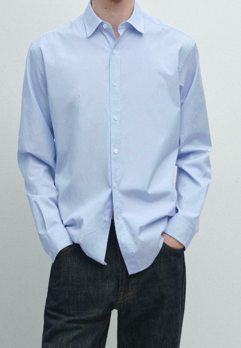 Homme portant une chemise rayée bleu clair à boutons, les mains dans les poches, associée à un jean foncé sur fond uni.