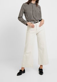 La blusa a maniche lunghe presenta un motivo geometrico in nero e crema, abbinata a pantaloni larghi crema a vita alta e mocassini neri.