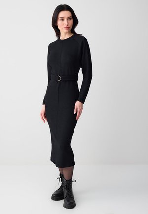 Kobieta w dopasowanej, czarnej, długorękawowej sukience midi z paskiem oraz czarnych, sznurowanych, wojskowych butach, stoi na tle gładkiego, jasnego tła.