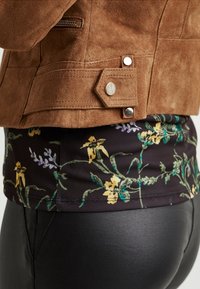 Chaqueta de ante marrón con acabado texturizado, que presenta botones plateados y una cintura con estampado floral en tela oscura con flores amarillas y verdes.