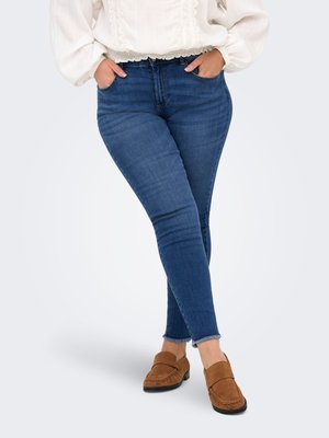 Kvinde iført blå skinny jeans med rå kant, hvid tekstureret langærmet bluse og brune ruskindsko, hænder i lommerne.