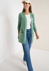 Zelený kardigan s otevřeným předním dílem, žebrovanými manžetami a dvěma předními kapsami, kombinovaný se světle modrými džíny a bílými teniskami.
