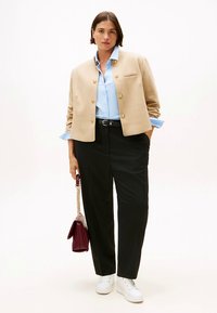 Beżowa marynarka ze złotymi guzikami na jasnoniebieskiej, w paski koszuli. Czarne spodnie i białe trampki. Trzyma bordową torebkę.