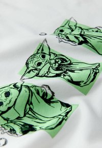Țesătură albă cu ilustrații repetate în negru și verde ale unui personaj cu urechi mari, ținând un obiect și purtând o robă.