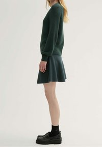 Suéter de punto verde con mangas largas, combinado con una falda oscura. Zapatos negros gruesos y calcetines completan el atuendo.