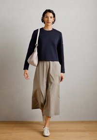 Mujer caminando en interiores con una blusa negra de manga larga, pantalones anchos beige, zapatillas blancas y llevando un bolso de hombro beige claro.