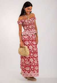 Robe à imprimé floral rouge et blanc, à bretelles dégagées, avec un corsage froncé, une taille peplum et une longue jupe. Le mannequin porte un sac en toile beige tissé.