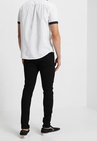 Camicia bianca a maniche corte con bordi neri, abbinata a jeans neri slim-fit e scarpe slip-on nere, vista da dietro in un contesto neutro.