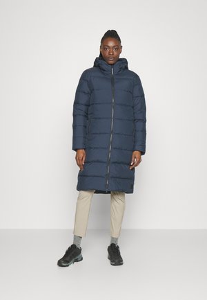 Marineblauwe pufferjas met capuchon, verticale stiknaden, volledige ritssluiting en zijzakken, gedragen over crème kleurige broek en zwarte sneakers.