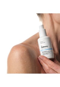 Hand holding a white bottle of The Ordinary Niacinamide 5% Face and Body Emulsion near a shoulder and neck against a white background.