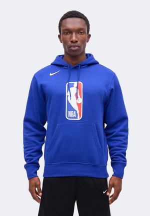 Muž, ktorý má na sebe modrú mikinu NBA s logom vpredu a čierne nohavice Nike, stojí čelom vpred na jednofarebnom pozadí.