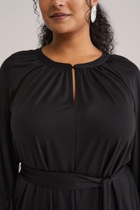 Schwarzes langärmeliges Kleid mit einem Schlüsselloch-Ausschnitt und gerafften Schultern. Es verfügt über eine gebundene Taille und eine glatte Textur. Silberne Ohrringe runden den Look ab.