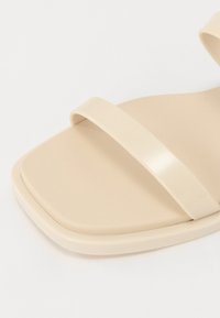 Beige plastik sandaler med to brede stropper og et kvadratisk tådesign. Mat tekstur og glat overflade, med en lav profil sål.