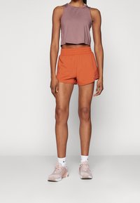 Top corto marrón deportivo, pantalones cortos deportivos naranjas con cintura elástica, combinados con zapatillas de deporte rosa claro y calcetines blancos.