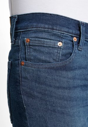 Gros plan d'un jean en denim bleu foncé montrant la zone de poche avant avec des rivets en cuivre et des détails de couture visibles.