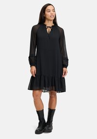 Das schwarze Kleid hat einen gestuften, rüschengeschmückten Saum, lange transparente Ärmel und einen Kordelzug-Ausschnitt. Kombiniert mit schwarzen Schnürstiefeln.