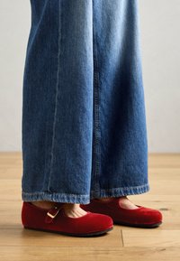 Zapatos Mary Jane de terciopelo rojo con una correa y hebilla, combinados con jeans de mezclilla azul de pierna ancha, sobre un suelo de madera.