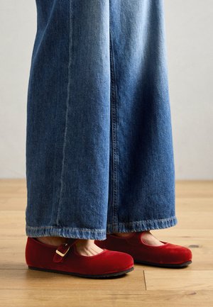 Røde fløyels Mary Jane-sko med stropp og spenne, kombinert med vide blå denimjeans, plassert på et treverk gulv.