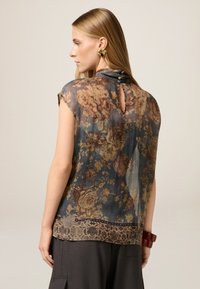 Durchscheinende Bluse mit floralen Mustern auf einem blau-grauen Grund, kurzen Ärmeln und einer Schlüsselaugenöffnung am Rücken, die mit Knöpfen gesichert ist. Akzentuiert mit einem dekorativen Saum.