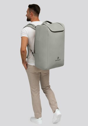 Un homme portant un t-shirt blanc, un pantalon beige et des baskets blanches porte un grand sac à dos rectangulaire gris avec le logo "pactastic".