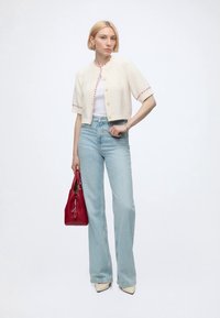 Femme aux cheveux blonds courts portant un cardigan court blanc boutonné, un jean taille haute bleu clair, des chaussures crème pointues et tenant un sac à main rouge.