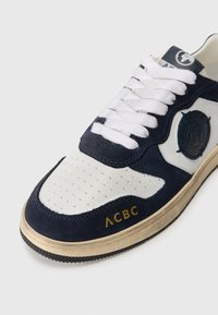 Námornícka semišová a biela kožená teniska s perforovanou špičkou, okrúhlymi šnúrkami a zlatým logom "ACBC" na boku. Gumená podrážka s textúrovaným detailom.
