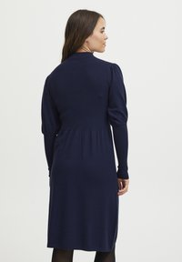 Mörkblå stickad klänning med långa ärmar, med puffade axlar, en åtsittande bodice och en utsvängd kjol som slutar under knät.