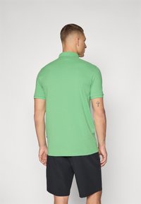 Grön bomullspolo med krage, korta ärmar och rak nederkant, bärs med svarta shorts. Modellen sedd bakifrån.