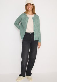 Cardigan texturé vert clair avec col rond, fermeture �à boutons, associé à un haut clair, un jean noir et des baskets beiges.