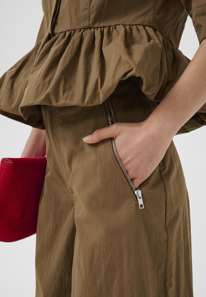 Persoon draagt bruine broek en bijpassend bovenstuk met een ritszak, houdt een kleine rode clutch met reliëf in de linkerhand.