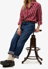 Camicia in flanella a quadri rossi e bianchi, jeans di denim, cintura intrecciata marrone, calzini color crema, scarpe scure. Seduto su uno sgabello di legno.