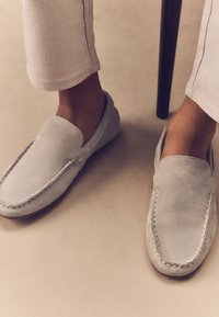 Gray suede slip-on shoes with stitched detailing, flat sole, and rounded toe, paired with light-colored, tapered pants in a neutral setting.