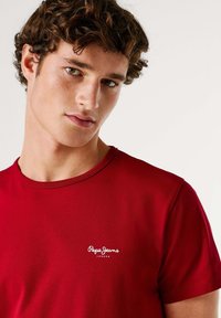 Rood katoenen T-shirt met een ronde hals. Het heeft een klein wit logo van "Pepe Jeans London" op het linkerbovenstuk van de borst. Glad oppervlak.