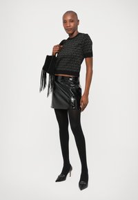 Maglione corto nero con motivo testurizzato, abbinato a una mini gonna in pelle nera lucida e collant neri; modella tiene in mano una borsa nera con frange.