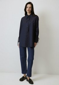 Camicia blu navy con colletto a papillon, maniche lunghe con laccetti ai polsi, abbinata a jeans in denim scuro e mocassini neri.