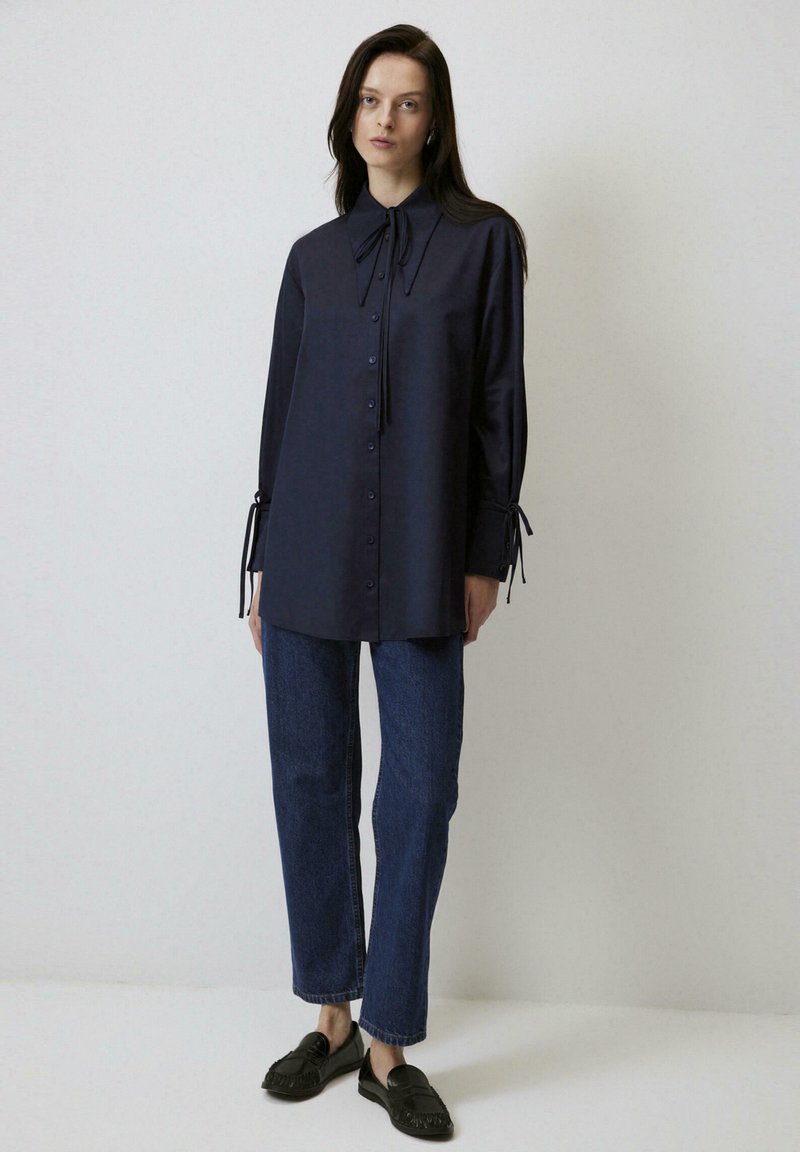 Camicia blu navy con colletto a papillon, maniche lunghe con laccetti ai polsi, abbinata a jeans in denim scuro e mocassini neri.