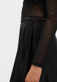 Fekete ruha sima, fényes textúrával és szűkített bodiccal. Hosszú, áttetsző ujjakkal rendelkezik, és diszkrét cipzárral az oldalon.