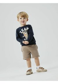 Sudadera de forro polar en color marino con la palabra "TEAM" en blanco y un dibujo de perro, pantalones cortos beige y zapatillas claras con detalles marrones. Mangas cortas y pantalones cortos con puños.