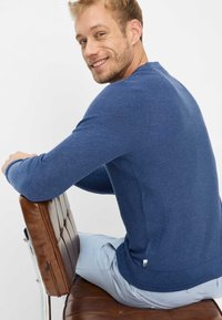 Blauer Strickpullover mit langen Ärmeln und gerippten Bündchen, getragen von einer Person, die auf einem braunen Lederstuhl sitzt, kombiniert mit hellen Hosen.