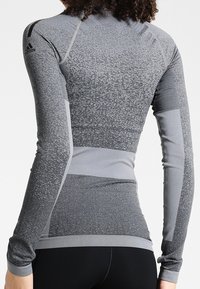 Dopasowana, długorękawowa bluzka sportowa w odcieniach szarości. Posiada teksturowany wzór z kontrastującymi, gładkimi akcentami w kolorze szarym, miękką tkaninę oraz kołnierz stojący.