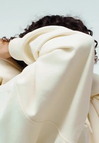 Krémová oversized mikina s mäkkou textúrou, rebrovanými manžetami a lemom; má voľný strih s objemnými rukávmi a bez viditeľných vzorov.