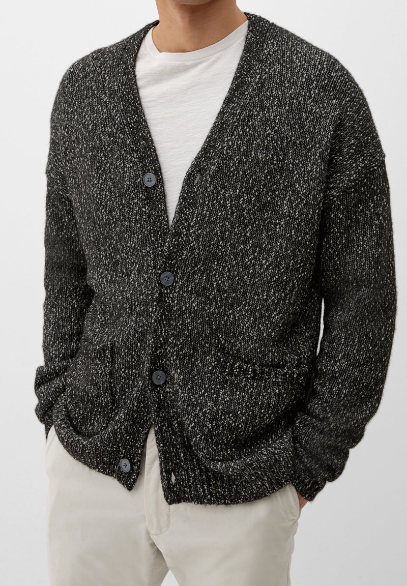Cardigan a maglia a pois bianchi e neri con scollatura a V, tasche frontali e cinque bottoni. Il materiale sembra avere una texture accogliente e morbida.