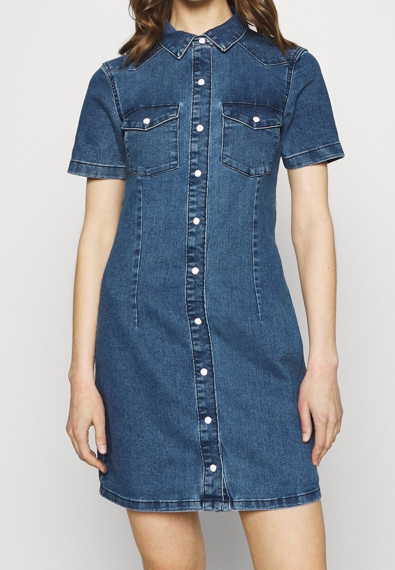 Femme portant une robe en denim bleu à manches courtes avec fermeture à boutons sur le devant et deux poches poitrine à rabat.