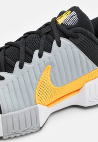 Scarpa sportiva nera con tomaia in mesh testurizzato, accenti in gomma grigia, un Swoosh arancione e suola bianca. Presenta elementi di design scanalati.