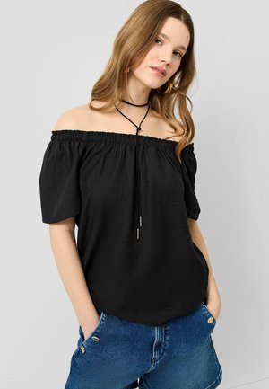 Jeune femme portant un haut noir épaules dénudées, un short en jean bleu, les mains dans les poches et un collier ras du cou noir sur un fond uni.