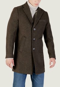 Cappotto in misto lana marrone con revers, tre grandi bottoni e tasche laterali, indossato sopra un maglione grigio chiaro, abbinato a jeans in denim.