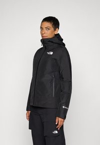 Zwarte waterdichte jas met capuchon, ritszakken aan de voorkant en het The North Face-logo op de borst en mouw. Glad textuur, aansluitend ontwerp.
