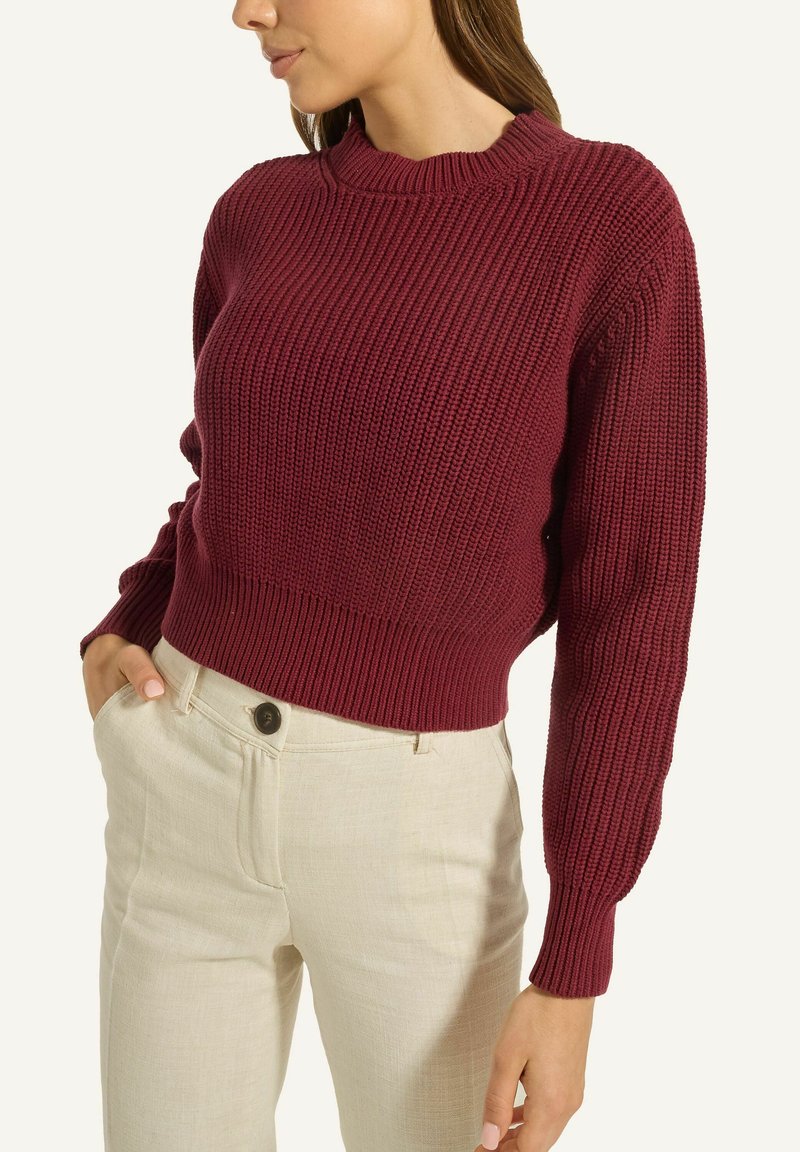 Frau trägt einen bordeauxroten, gestrickten Pullover mit langen Ärmeln und cremefarbene, hochtaillierte Hosen mit Knopfverschluss.