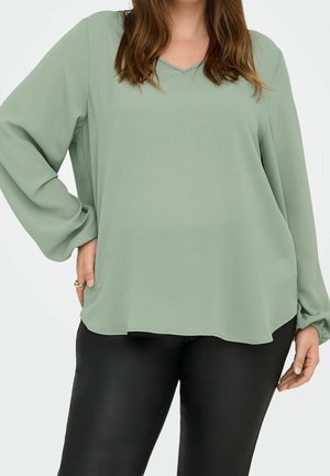 Frau trägt ein locker sitzendes, salbeigrünes Langarmshirt mit V-Ausschnitt und schwarze Hose, steht mit einer Hand in der Hüfte.